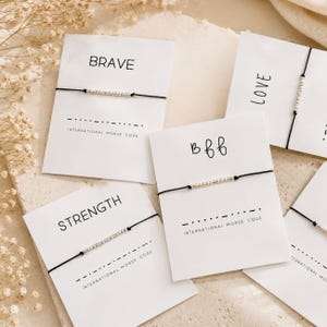 Può includere: Diversi braccialetti in cordino nero con piccole perline metalliche disposte in sequenze di codice Morse su cartoncini bianchi. Ogni cartoncino riporta una parola specifica come BRAVE, LOVE, STRENGTH o BFF, insieme alla sequenza in codice Morse.