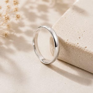 Pode incluir: Um anel de banda de cor prateada polida com uma superfície exterior lisa e arredondada e um design simples e limpo. O metal reflete a luz, criando um brilho intenso ao longo da borda curva da banda circular que se encontra na vertical.