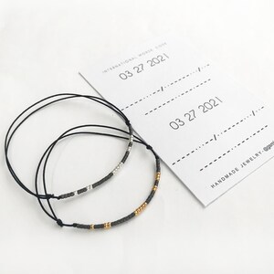 Puede incluir: Dos pulseras de cuerda negra con cuentas plateadas y doradas. Las pulseras están sobre un fondo blanco con una tarjeta que dice "Día Internacional de la Mujer 03 27 2021" y "Joyería hecha a mano - @gemminimal".