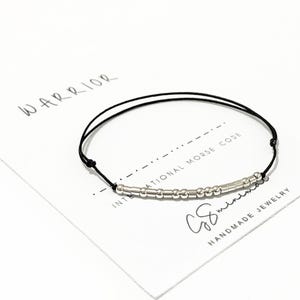 Könnte beinhalten: Ein schwarzes Armband aus Schnur mit silbernen Perlen, die "WARRIOR" im internationalen Morsecode buchstabieren. Das Armband liegt auf einer weißen Karte mit dem Text "WARRIOR" und "INTERNATIONAL MORSE CODE", der in schwarzer Tinte geschrieben ist. Die Karte trägt auch den Text "C&S handmade jewelry", der in schwarzer Tinte geschrieben ist.