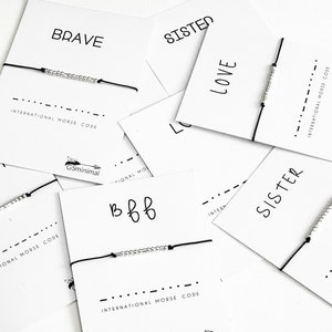 Op de afbeelding: Een verzameling armbanden met zwart touw en zilveren kralen. Elke armband heeft een woord op een kaart, waaronder "Brave", "Sister", "Love" en "Bbb". De kaarten bevatten ook de tekst "Internationale Morsecode".