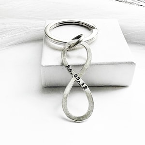 Könnte beinhalten: Silberner Schlüsselanhänger in Form eines Unendlichkeitszeichens mit einem eingravierten Datum. Der Schlüsselanhänger liegt auf einer weißen Oberfläche.