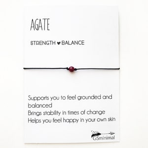 Peut inclure: Un bracelet en cordon noir avec une perle d'agate rouge foncé. La carte indique "AGATE", "STRENGTH BALANCE", "Supports you to feel grounded and balanced", "Brings stability in times of change", "Helps you feel happy in your own skin", et "GSminimal".