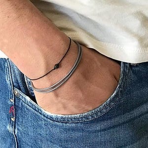 May include: A person wearing a black cord bracelet with a small black bead and a silver chain bracelet.
