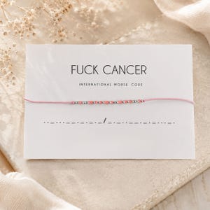Puede incluir: Una pulsera de hilo rosa con pequeñas cuentas plateadas y rosas dispuestas en código Morse, presentada en una tarjeta blanca con el texto FUCK CANCER INTERNATIONAL MORSE CODE y una secuencia de código Morse impresa bajo la pulsera.