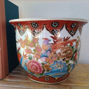 May include: A ceramic planter featuring a white base with a colorful hand-painted design of birds, pink and blue flowers, and gold accents. The rim and base are decorated with a scalloped pattern in deep red, black, and gold, creating a detailed finish.