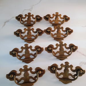 May include: A set of six brass-toned metal drawer pulls arranged in three rows of two. Each pull features a decorative backplate with a central pineapple motif and a curved, swinging handle attached to two rounded mounting points on the sides.