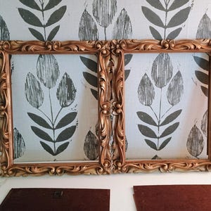 May include: Two rectangular picture frames with ornate, carved gold-toned borders featuring scrolling leaf and floral motifs. The frames are empty, showcasing a hollow center, and are accompanied by two separate dark brown backing boards.