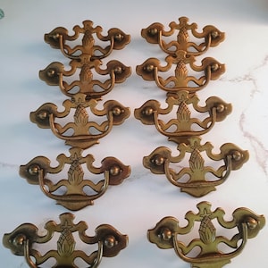 May include: A set of ten brass drawer pulls arranged in five rows of two. Each pull features a decorative backplate with a central pineapple motif and a curved, swinging handle attached to two side mounting points with rounded ends.