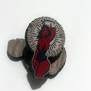 May include: A wooden pin with a red and black illustration of a pin-up style woman with horns and the word "Sinner" above her. The background is a white and gray sunburst pattern.