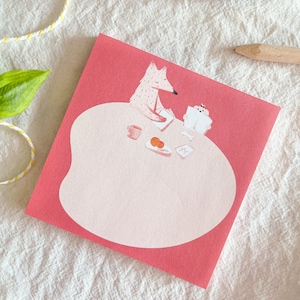 Pode incluir: Um bloco de notas rosa e vermelho com uma raposa e um animal branco sentados à mesa com uma xícara de chá, um prato de comida e um livro. O bloco de notas tem uma área rosa de forma irregular para escrever.