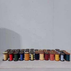 Puede incluir: Una colección de carretes de madera vintage llenos de hilos coloridos. Los carretes están dispuestos en filas, mostrando una variedad de colores que incluyen rojo, azul, amarillo, verde y marrón. Los carretes son de diferentes alturas y diámetros.