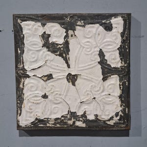 Puede incluir: Baldosa de techo cuadrada de metal antiguo con un acabado de pintura blanca descascarillada. La baldosa presenta un patrón floral repetitivo dentro de un borde gris oscuro. La pintura está desconchada, revelando el metal debajo, dándole un aspecto envejecido.