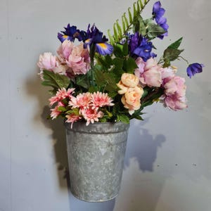 Puede incluir: Un cubo de metal galvanizado lleno de una variedad de flores artificiales coloridas. El arreglo incluye margaritas rosas, rosas melocotón, iris moradas y peonías rosas. El cubo está colgado en una pared gris claro.