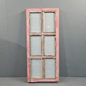 Può includere: Una cornice di finestra rettangolare vintage con sei vetri. La cornice è dipinta di rosa anticato, con usura visibile e vernice scheggiata. I vetri sono trasparenti. Sfondo grigio.