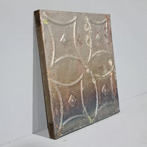 May include: A silver metal ceiling tile with a raised embossed pattern of overlapping circles and diamond shapes. The tile has a distressed, aged look with some areas of rust and discolouration.