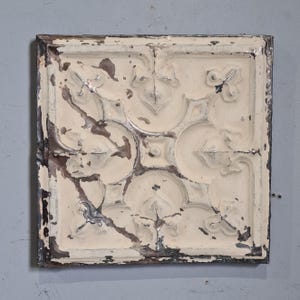 Puede incluir: Baldosa decorativa cuadrada de metal blanquecino con un patrón floral en relieve. La baldosa tiene un aspecto envejecido y desgastado con detalles en marrón oscuro y plateado en los bordes y dentro del diseño. Para la decoración del hogar.