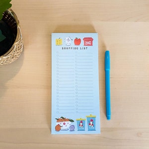 May include: A blue notepad with a shopping list on it. The notepad has a cute design with illustrations of fruit, a bowl, and jars. The text "SHOPPING LIST" is printed on the notepad. A blue pen is lying next to the notepad.