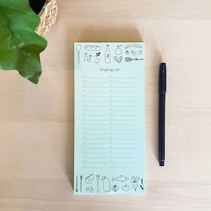 Op de afbeelding: Een lichtblauw notitieboekje met een kopje "Boodschappenlijstje" en een checklist. Het notitieboekje is versierd met zwart-witte illustraties van voedingsmiddelen, waaronder een brood, een fles wijn, een peer en een avocado.