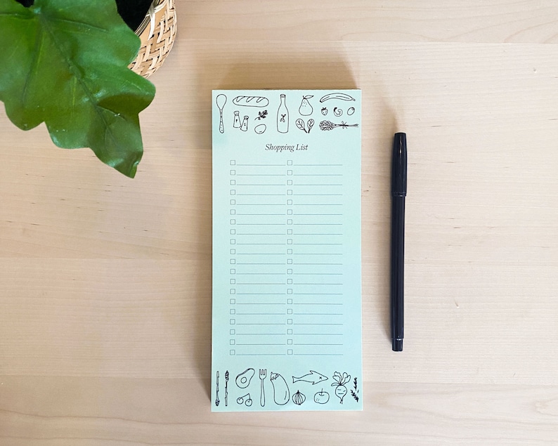 Op de afbeelding: Een lichtblauw notitieboekje met een kopje "Boodschappenlijstje" en een checklist. Het notitieboekje is versierd met zwart-witte illustraties van voedingsmiddelen, waaronder een brood, een fles wijn, een peer en een avocado.