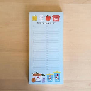 May include: A blue notepad with a shopping list on it. The notepad has illustrations of a teacup, an apple, a box of sugar, and a box of flour. The text "SHOPPING LIST" is printed on the notepad.