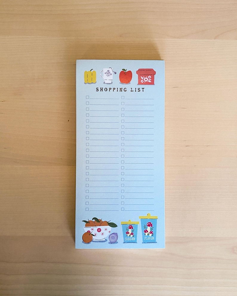 May include: A blue notepad with a shopping list on it. The notepad has illustrations of a teacup, an apple, a box of sugar, and a box of flour. The text "SHOPPING LIST" is printed on the notepad.