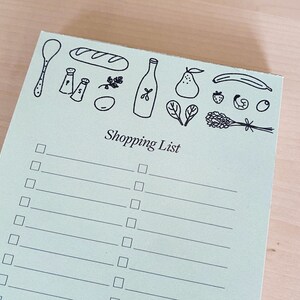 Op de afbeelding: Een groene boodschappenlijst met een zwart-witte illustratie van een brood, een fles olie, een peer, een banaan, een aardbei, een lavendelbosje en een lepel. Het notitieblok heeft een titel die "Shopping List" zegt en heeft 15 lege regels om op te schrijven.