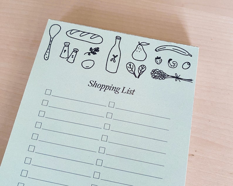 Op de afbeelding: Een groene boodschappenlijst met een zwart-witte illustratie van een brood, een fles olie, een peer, een banaan, een aardbei, een lavendelbosje en een lepel. Het notitieblok heeft een titel die "Shopping List" zegt en heeft 15 lege regels om op te schrijven.