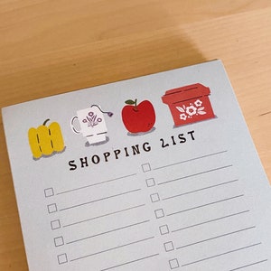May include: A blue shopping list notepad with a red apple, a yellow pepper, a white mug, and a red box with white flowers. The notepad has the words "SHOPPING LIST" printed on it.