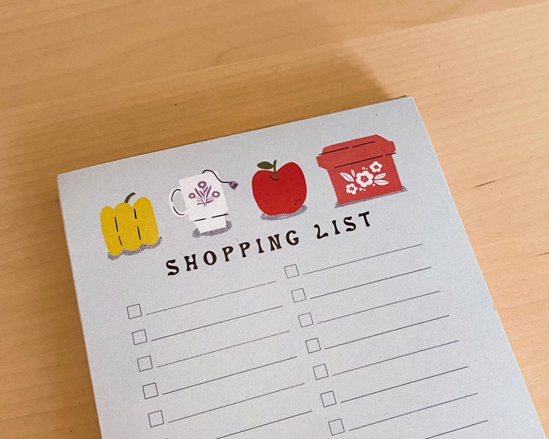 May include: A blue shopping list notepad with a red apple, a yellow pepper, a white mug, and a red box with white flowers. The notepad has the words "SHOPPING LIST" printed on it.