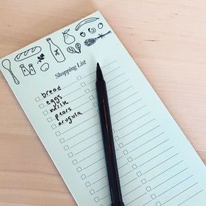 Op de afbeelding: Een lichtblauw notitieboekje voor boodschappen met een zwarte pen erop. Het notitieboekje heeft een checklist met items zoals brood, eieren, melk, peren en rucola erop geschreven. Bovenaan het notitieboekje staat een grillige illustratie van een brood, een fles wijn, een peer, een tros druiven en andere etenswaren.