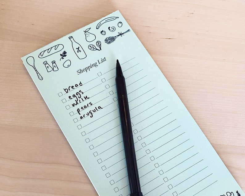 Op de afbeelding: Een lichtblauw notitieboekje voor boodschappen met een zwarte pen erop. Het notitieboekje heeft een checklist met items zoals brood, eieren, melk, peren en rucola erop geschreven. Bovenaan het notitieboekje staat een grillige illustratie van een brood, een fles wijn, een peer, een tros druiven en andere etenswaren.