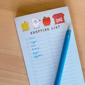 May include: A blue shopping list notepad with a checklist and handwritten items including flour, sugar, tea, bread, eggs, and apples. The notepad has a red apple, a yellow pepper, a white mug, and a red container with flowers on the top.