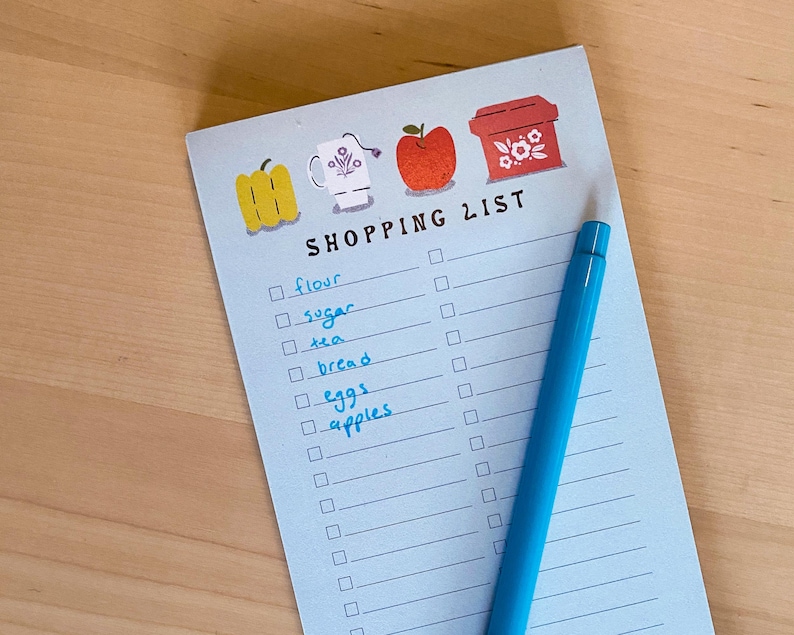 May include: A blue shopping list notepad with a checklist and handwritten items including flour, sugar, tea, bread, eggs, and apples. The notepad has a red apple, a yellow pepper, a white mug, and a red container with flowers on the top.