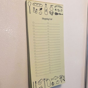 Op de afbeelding: Een geel notitieboekje met een boodschappenlijstje erop. Het notitieboekje heeft een zwarte rand en is versierd met tekeningen van etenswaren, waaronder een stokbrood, een fles wijn, een peer, een banaan, een aardbei, een tros druiven, een takje peterselie, een bos spinazie, een bos lavendel, een lepel, een vork, een mes, een bos asperges, een avocado, een aubergine, een ui, een appel, een radijs en een vis.