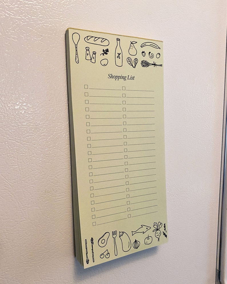 Op de afbeelding: Een geel notitieboekje met een boodschappenlijstje erop. Het notitieboekje heeft een zwarte rand en is versierd met tekeningen van etenswaren, waaronder een stokbrood, een fles wijn, een peer, een banaan, een aardbei, een tros druiven, een takje peterselie, een bos spinazie, een bos lavendel, een lepel, een vork, een mes, een bos asperges, een avocado, een aubergine, een ui, een appel, een radijs en een vis.