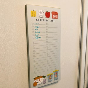 May include: A blue and white shopping list notepad with a checklist and illustrations of a yellow pepper, a white teacup, a red apple, and a red box with white flowers. The notepad has the words "SHOPPING LIST" printed at the top. The checklist includes items such as "flour", "sugar", "tea", "bread", "eggs", and "apples".