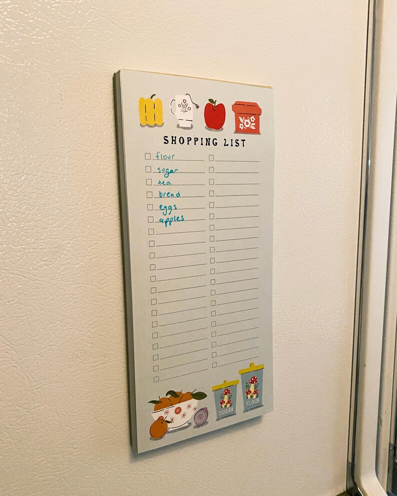 May include: A blue and white shopping list notepad with a checklist and illustrations of a yellow pepper, a white teacup, a red apple, and a red box with white flowers. The notepad has the words "SHOPPING LIST" printed at the top. The checklist includes items such as "flour", "sugar", "tea", "bread", "eggs", and "apples".