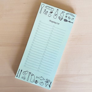 Op de afbeelding: Een lichtblauw notitieblok voor boodschappen met een zwart-witte illustratie van verschillende boodschappen bovenaan en onderaan. De tekst "Shopping List" staat in het zwart bovenaan het notitieblok.