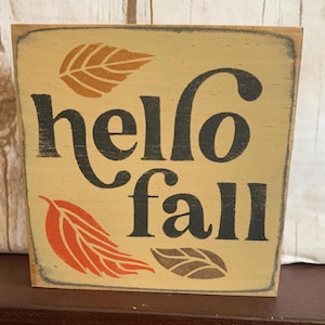 May include: A square wooden sign with the words "hello fall" in black script. The sign has a light beige background with orange and brown leaf designs. The edges of the sign are distressed.