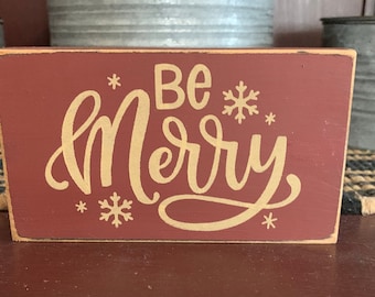 May include: A rectangular, maroon wooden sign with the words "Be Merry" in gold script and snowflake accents. The sign has a distressed, rustic look, perfect for holiday decor.