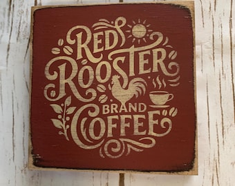 May include: A square, rustic wooden sign with a distressed red background. The sign features the words "Red Rooster Brand Coffee" in a decorative, cream-colored font, along with a rooster illustration, coffee beans, and a steaming coffee cup.