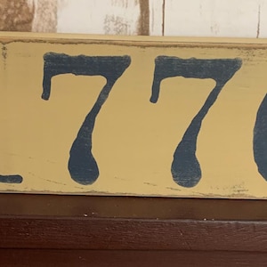 May include: A wooden sign with the number 1776 painted in blue on a yellow background. Two red stars are on either side of the number.
