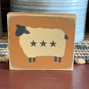 May include: A wooden block with a painted image of a white sheep with three black stars on its back. The sheep is standing on a brown background.