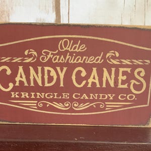May include: A rectangular wooden sign with a burgundy background and gold lettering. The sign reads "Olde Fashioned CANDY CANES" with "KRINGLE CANDY CO." below. The sign has a distressed, vintage look.