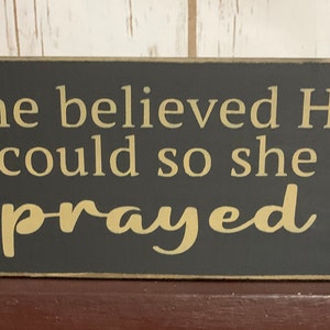 May include: A dark gray wooden sign with the text "She believed He could so she prayed" in a light gold color.