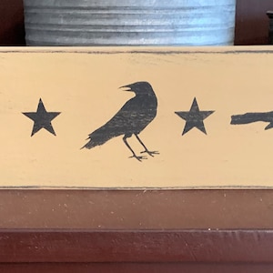 May include: A wooden sign with a distressed, yellow paint finish. Three black crow silhouettes are spaced evenly across the sign, with a black star between each crow.