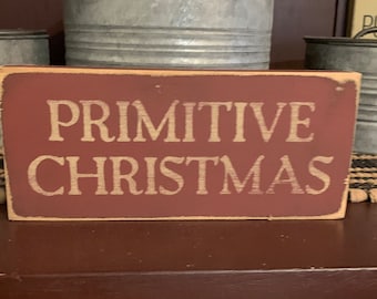 May include: A red wooden sign with white lettering that reads "Primitive Christmas".