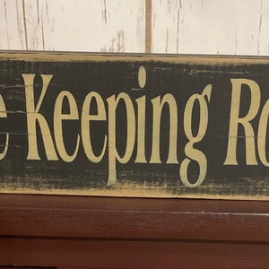 May include: A black wooden sign with white distressed paint and the text "The Keeping Room" in gold lettering.