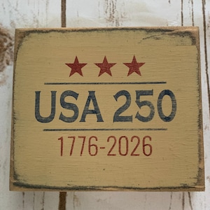 May include: A rectangular wooden sign with a distressed finish. The sign features the text "USA 250" in navy blue, with three red stars above and the years "1776-2026" in red below. The background is a light tan color.